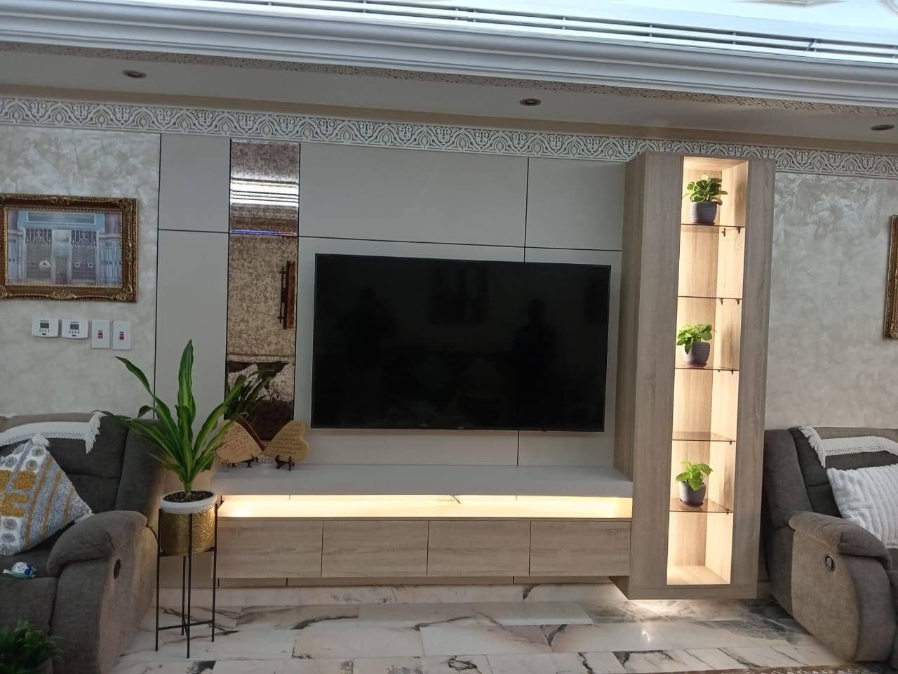 مجلس عائلي بوحدة تلفاز خشبية وخزائن بيضاء ورفوف زجاجية مضاءة وإضاءة مخفية أسفل الوحدة