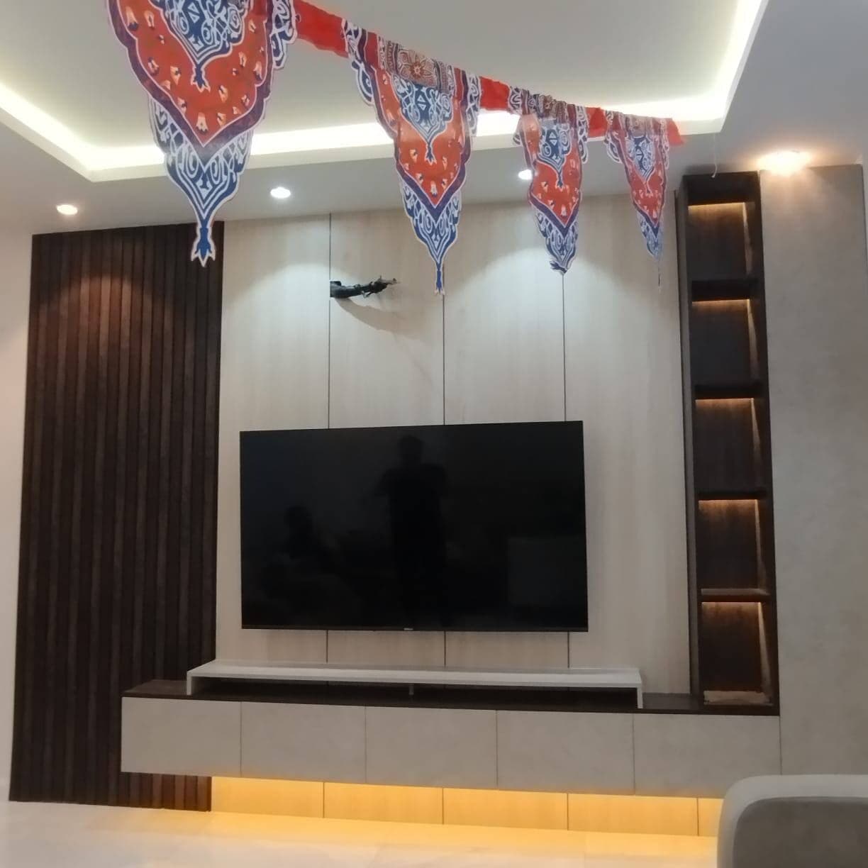 جدار تلفاز بألواح فاتحة يحده عمود خشب مخدد داكن من جهة وعمود رفوف بإضاءة خفية من الجهة الأخرى، فوق وحدة سفلية معلقة بإضاءة سفلية دافئة