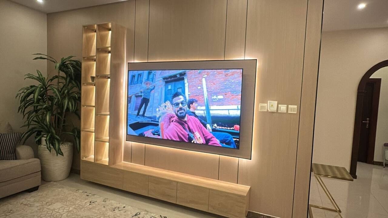 جدار مكسو بخشب فاتح بألواح رأسية، بشاشة يحيط بها إطار بإضاءة دافئة خفية، وعمود رفوف زجاجية مضاءة، فوق وحدة سفلية ممتدة بجانب كنبة ونبتة